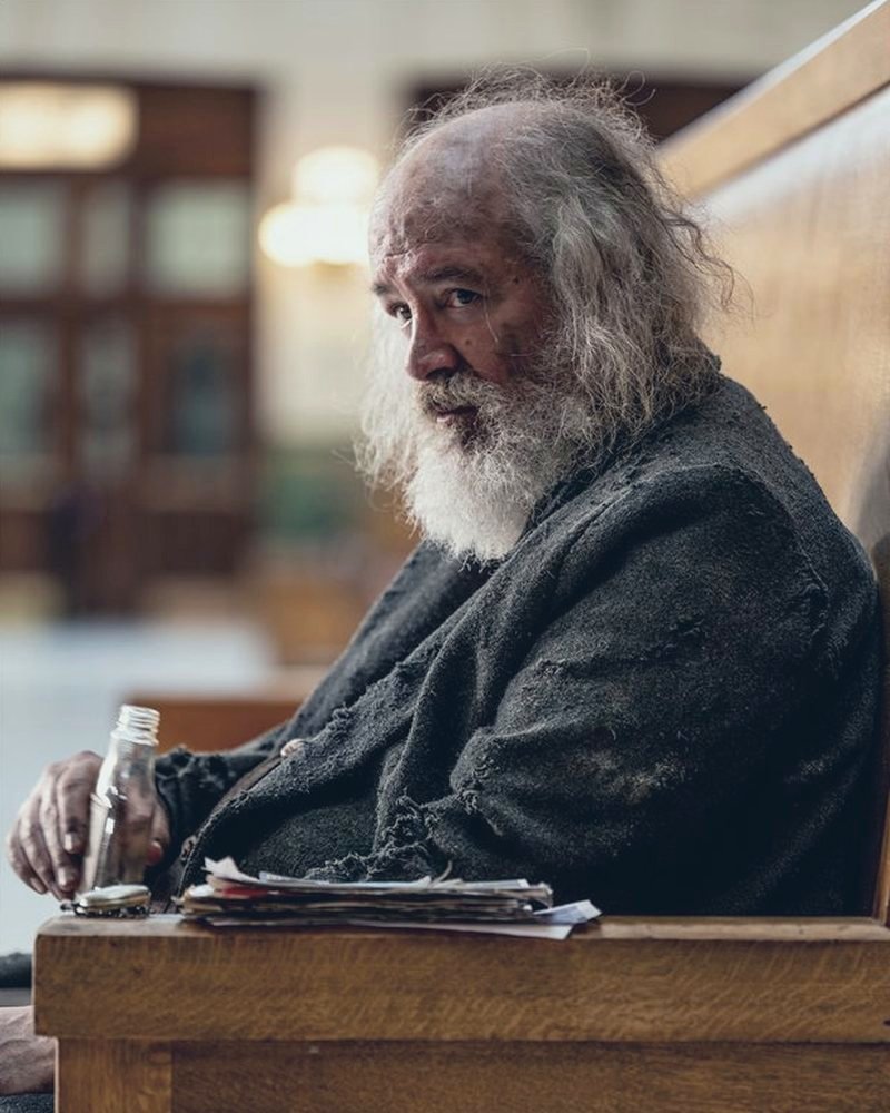 Arthur sitting on a wooden station bench with a bottle and a stack of tickets, looking back over his shoulder