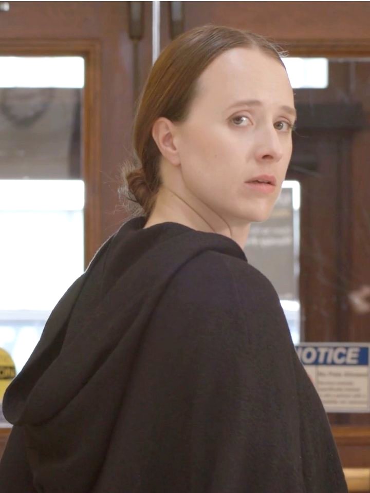 Geena Pietromonaco as Margaret, in a dark hooded cloak, looking back over her shoulder